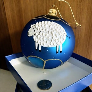 May include: A spherical blue glass Christmas ornament featuring a raised white textured sheep illustration. The ornament has a gold hanging loop and is displayed in a blue box with a small circular coin placed on the white interior base.