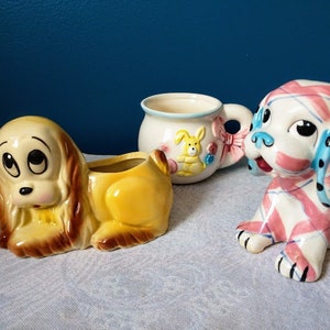 May include: Three ceramic dog figurines and a mug. The dog figurines are in various poses and colors, including a yellow and brown planter, and a white dog with pink and blue stripes. The mug is white with a bunny design.