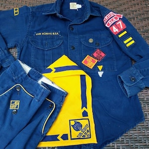 May include: A blue Cub Scout uniform with a yellow neckerchief, patches, and the text "Cub Scouts B.S.A." on the shirt. The uniform includes a blue shirt, pants, and a neckerchief.