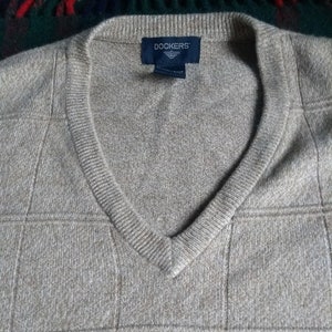 May include: A tan V-neck sweater with a grid pattern. The sweater has a ribbed neckline and the Dockers brand label is visible. The fabric appears to be a soft knit.