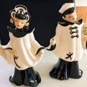 May include: Two ceramic figurines depicting figures in white and black outfits. The left figure wears a white headpiece and holds a yellow floral fan. The right figure wears a white cap and holds a yellow floral bouquet. Both have black skirts.