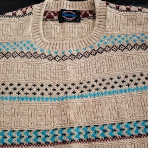 May include: A beige crew neck sweater featuring a textured knit pattern and horizontal bands of geometric designs in bright blue, dark maroon, and black. The interior collar tag displays the brand name Jantzen and the text Made in USA.