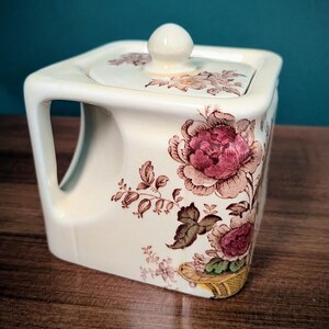 May include: A square ceramic sugar bowl with a cream-colored base and a matching lid featuring a knob handle. The exterior is decorated with a detailed transfer print of pink and brown floral patterns, including large peony-like blossoms.