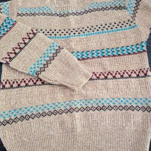 May include: A beige long-sleeve crew neck sweater featuring a textured knit pattern with horizontal bands of geometric designs in bright blue and dark maroon. The sweater has ribbed cuffs, a ribbed hem, and a ribbed neckline with a small interior label.