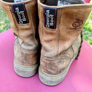 May include: A pair of tan leather work boots featuring a dark brown fabric pull tab with the text Georgia Boot in white. The interior collar is lined with a dark brown mesh material, and a white label with black text is visible inside the boot.
