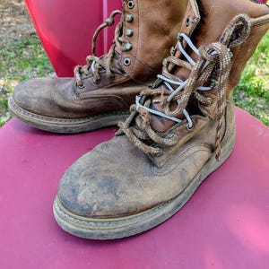 May include: A pair of worn, tan leather work boots featuring a high-top design, rounded toes, and thick, textured brown laces. The boots have metal eyelets and hooks for lacing, with visible scuff marks on the toes and a sturdy, flat rubber sole.