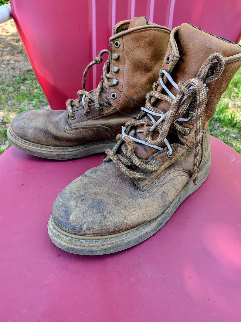 May include: A pair of worn, tan leather work boots featuring a high-top design, rounded toes, and thick, textured brown laces. The boots have metal eyelets and hooks for lacing, with visible scuff marks on the toes and a sturdy, flat rubber sole.