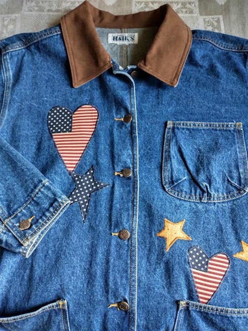 May include: A blue denim jacket with a brown corduroy collar. The jacket has several patches sewn on, including hearts, stars, and a star-spangled banner design. The text "Hanks" is visible on the label.