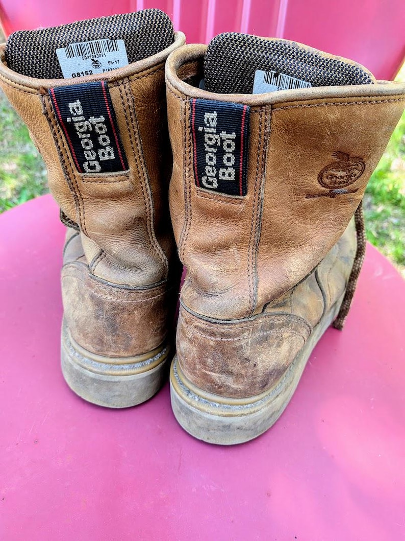 May include: A pair of tan leather work boots featuring a dark brown fabric pull tab with the text Georgia Boot in white. The interior collar is lined with a dark brown mesh material, and a white label with black text is visible inside the boot.