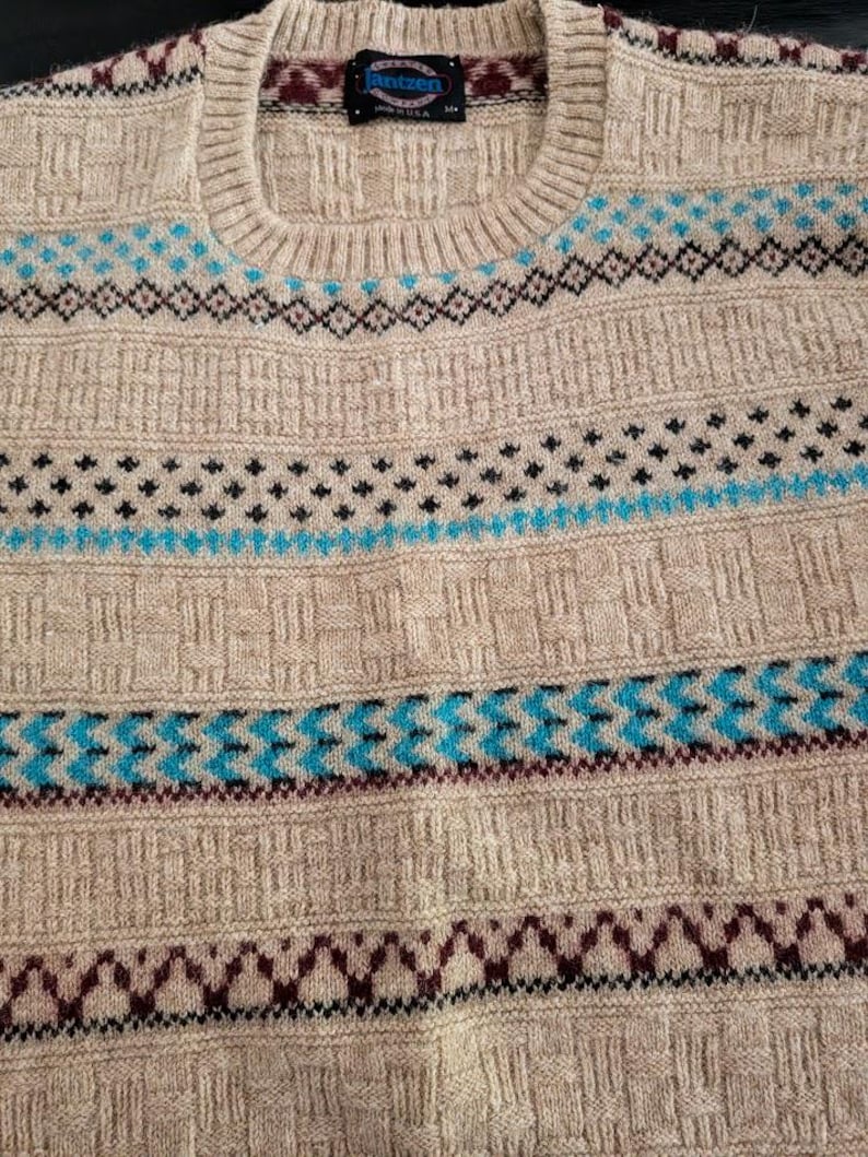 May include: A beige crew neck sweater featuring a textured knit pattern and horizontal bands of geometric designs in bright blue, dark maroon, and black. The interior collar tag displays the brand name Jantzen and the text Made in USA.