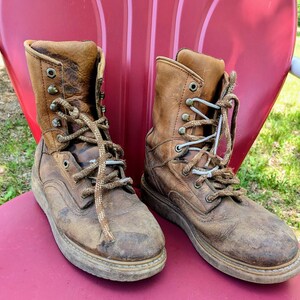 May include: A pair of worn, tan leather lace-up boots featuring a high-top design, metal eyelets, and thick, textured brown laces. The boots exhibit visible signs of wear, including scuffs and creases on the leather surface and sturdy soles.