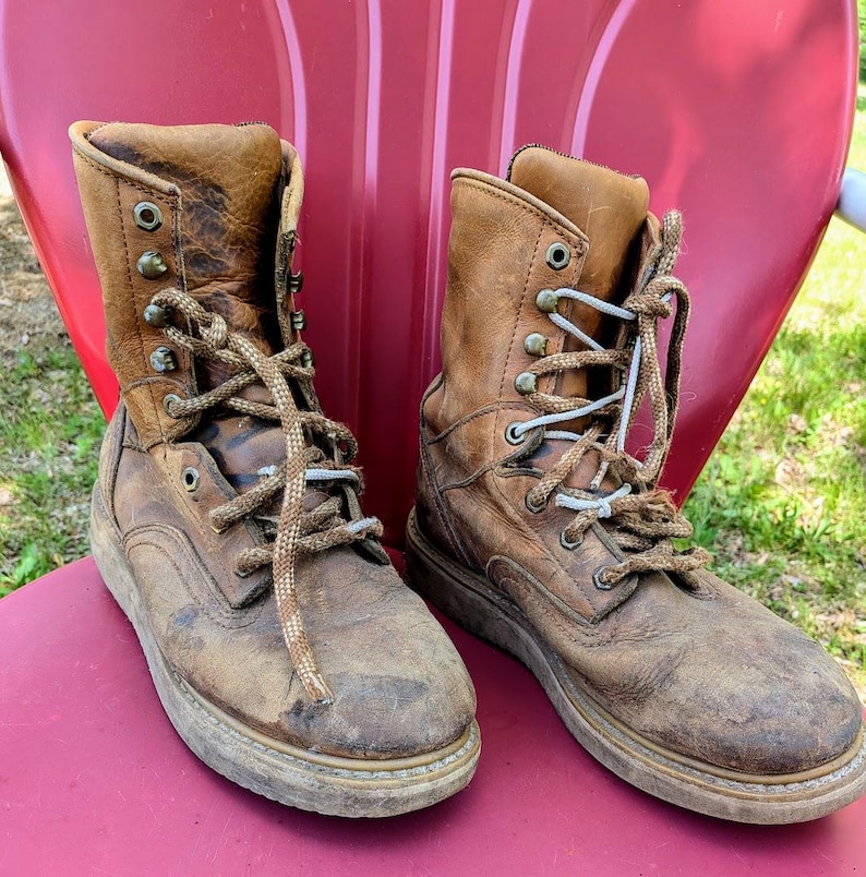 May include: A pair of worn, tan leather lace-up boots featuring a high-top design, metal eyelets, and thick, textured brown laces. The boots exhibit visible signs of wear, including scuffs and creases on the leather surface and sturdy soles.