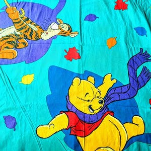 May include: A teal-colored bedsheet featuring cartoon illustrations of Winnie the Pooh and Tigger. Winnie the Pooh is yellow, wearing a red shirt and purple scarf. Tigger is orange with black stripes. Colorful leaves are scattered across the sheet.