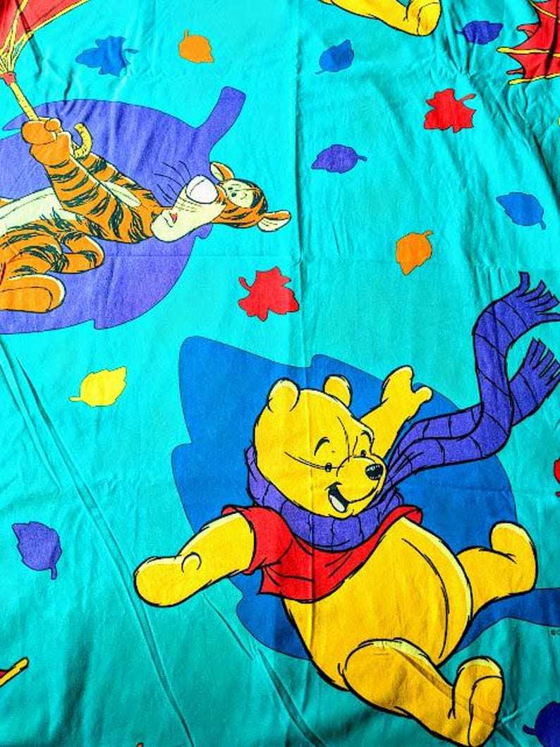 May include: A teal-colored bedsheet featuring cartoon illustrations of Winnie the Pooh and Tigger. Winnie the Pooh is yellow, wearing a red shirt and purple scarf. Tigger is orange with black stripes. Colorful leaves are scattered across the sheet.