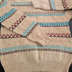 May include: A beige long-sleeve sweater featuring a textured knit pattern with horizontal bands of geometric designs in shades of maroon, black, and bright blue. The garment has a crew neckline and ribbed cuffs and hem, showcasing a detailed knit.