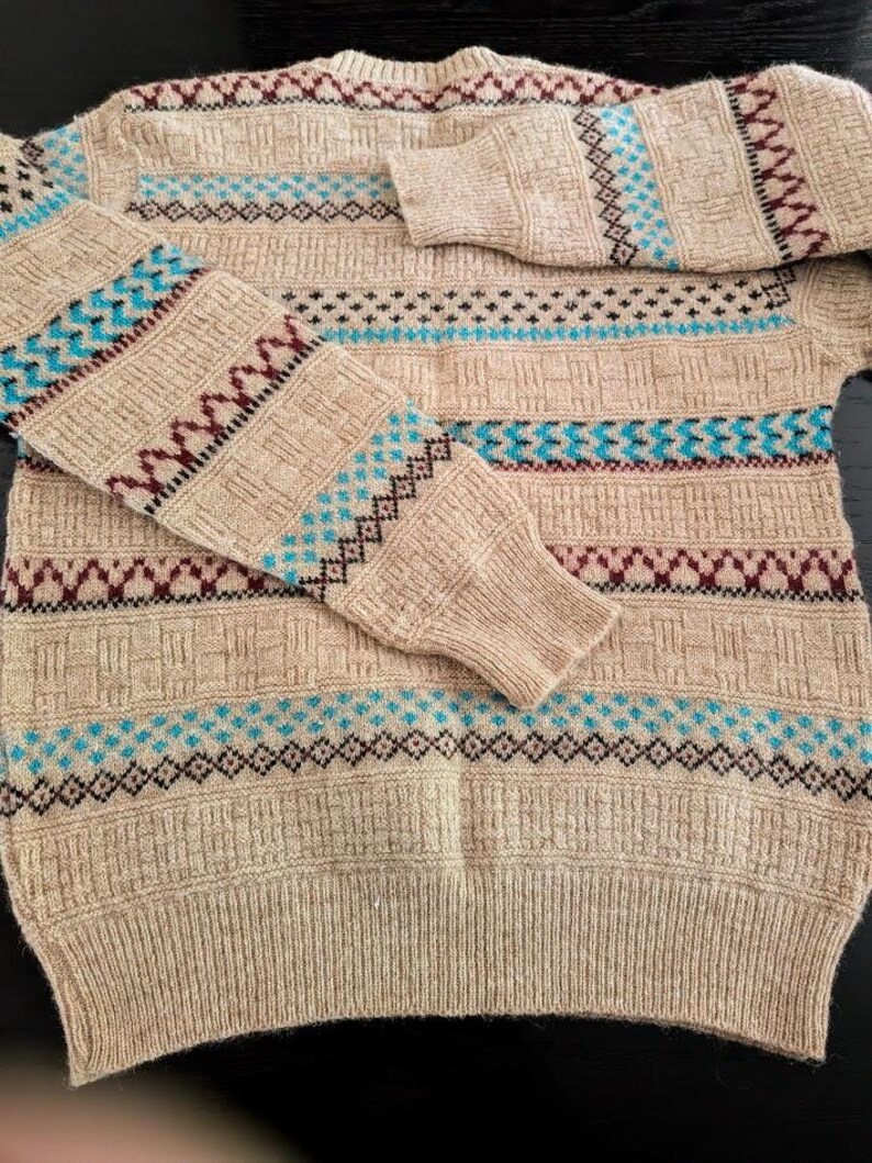 May include: A beige long-sleeve sweater featuring a textured knit pattern with horizontal bands of geometric designs in shades of maroon, black, and bright blue. The garment has a crew neckline and ribbed cuffs and hem, showcasing a detailed knit.