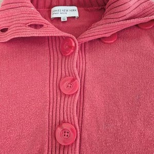 May include: A close-up of a coral-colored sweater with a ribbed collar and button-up front. The sweater has large, round buttons and a label that reads "JONES NEW YORK SPORT PETITE". The knitwear is a solid color.