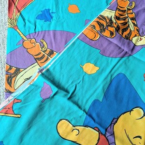 May include: A teal-colored bedsheet featuring cartoon illustrations of Winnie the Pooh and Tigger. The design includes purple, orange, and blue accents, with leaf and umbrella motifs. The sheet is folded, showing the pattern.