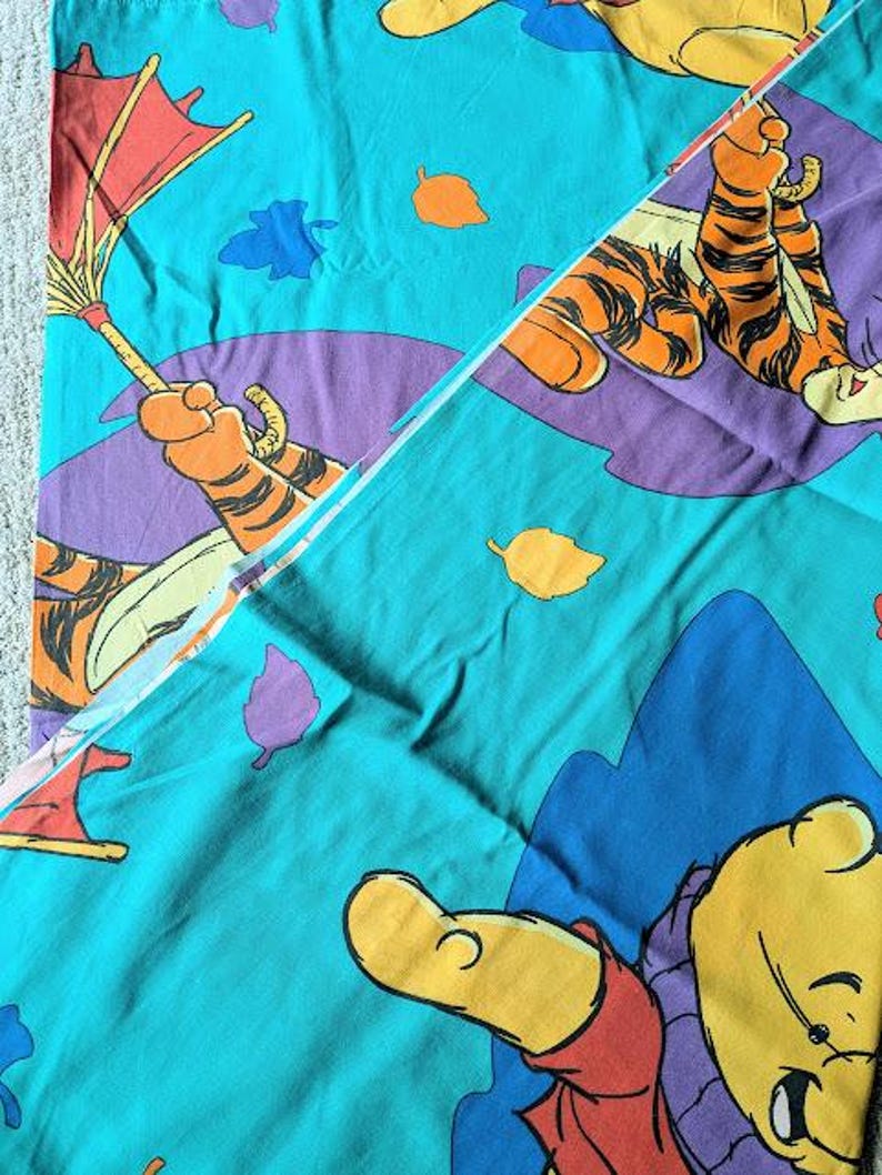 May include: A teal-colored bedsheet featuring cartoon illustrations of Winnie the Pooh and Tigger. The design includes purple, orange, and blue accents, with leaf and umbrella motifs. The sheet is folded, showing the pattern.