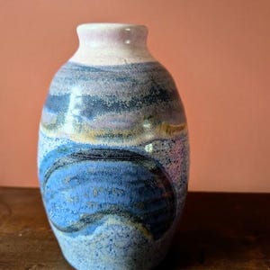May include: A handmade ceramic vase with a rounded body and a narrow neck. The vase features a blue, purple, and tan color scheme with a textured surface. The design includes abstract blue shapes. The vase is a decorative item.
