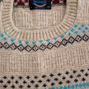 May include: A beige knit sweater featuring a crew neckline and a black label reading Jantzen Sweater Company Made in U.S.A. M. The sweater displays a textured pattern with horizontal bands of geometric shapes in maroon, black, and bright blue.