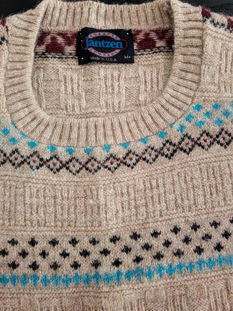 May include: A beige knit sweater featuring a crew neckline and a black label reading Jantzen Sweater Company Made in U.S.A. M. The sweater displays a textured pattern with horizontal bands of geometric shapes in maroon, black, and bright blue.