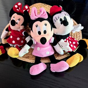 May include: Three plush Minnie Mouse dolls arranged together. The left doll wears a red dress with white polka dots and a matching bow. The centre doll wears a pink dress with white polka dots and a pink bow. The right doll wears a red dress with white polka dots and a red hat.