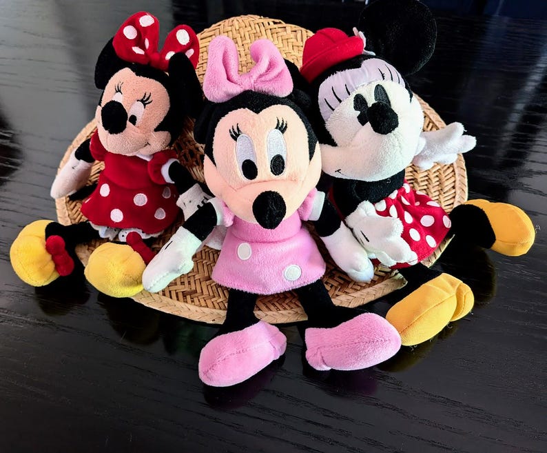 May include: Three plush Minnie Mouse dolls arranged together. The left doll wears a red dress with white polka dots and a matching bow. The centre doll wears a pink dress with white polka dots and a pink bow. The right doll wears a red dress with white polka dots and a red hat.