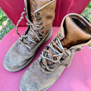 May include: A pair of worn, tan leather lace-up boots featuring a high-top design, rounded toes, and thick, textured brown laces. The boots have visible scuffs and signs of wear on the leather surface, metal eyelets, and a sturdy construction.