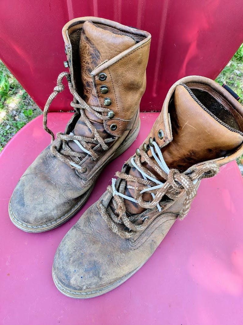 May include: A pair of worn, tan leather lace-up boots featuring a high-top design, rounded toes, and thick, textured brown laces. The boots have visible scuffs and signs of wear on the leather surface, metal eyelets, and a sturdy construction.