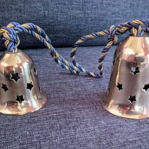 K&ouml;nnte beinhalten: Zwei silberfarbene Glocken mit sternf&ouml;rmigen Aussparungen, verbunden durch ein blau-goldenes gedrehtes Seil. Die Glocken stehen auf einer strukturierten blauen Oberfl&auml;che. Die Glocken haben ein gl&auml;nzendes Finish.