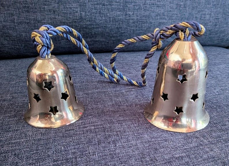 K&ouml;nnte beinhalten: Zwei silberfarbene Glocken mit sternf&ouml;rmigen Aussparungen, verbunden durch ein blau-goldenes gedrehtes Seil. Die Glocken stehen auf einer strukturierten blauen Oberfl&auml;che. Die Glocken haben ein gl&auml;nzendes Finish.