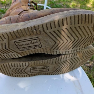 May include: A pair of tan leather work boots with brown laces and light-colored rubber soles. The soles feature a chevron tread pattern and a rectangular stamp that reads Georgia Boot Oil & Slip Resistant in the center of the bottom surface.