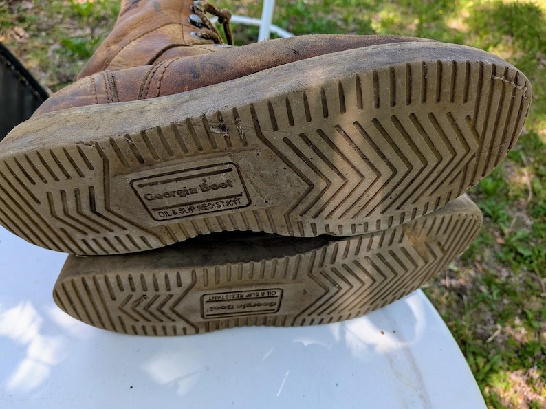 May include: A pair of tan leather work boots with brown laces and light-colored rubber soles. The soles feature a chevron tread pattern and a rectangular stamp that reads Georgia Boot Oil & Slip Resistant in the center of the bottom surface.