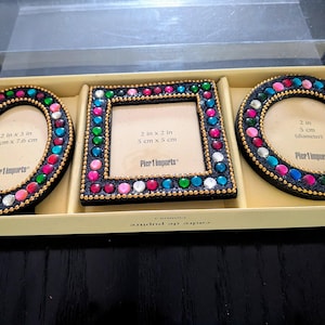 May include: A set of three decorative picture frames in a box. The frames are oval, square, and round, each with a black border and colorful gem accents. The oval frame is 2 in x 3 in (5 cm x 7.6 cm), the square frame is 2 in x 2 in (5 cm x 5 cm), and the round frame is 2 in (5 cm) in diameter.