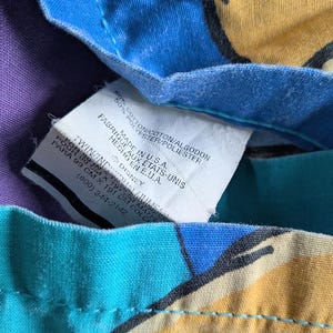 May include: Close-up of a tag on a colorful fabric. The tag reads "Made in U.S.A." and includes the Disney logo. The fabric has a purple, blue, teal, and yellow pattern. The tag also includes the dimensions of the fabric.