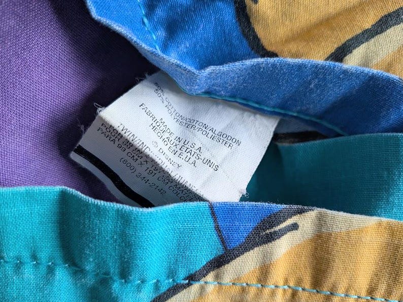 May include: Close-up of a tag on a colorful fabric. The tag reads "Made in U.S.A." and includes the Disney logo. The fabric has a purple, blue, teal, and yellow pattern. The tag also includes the dimensions of the fabric.