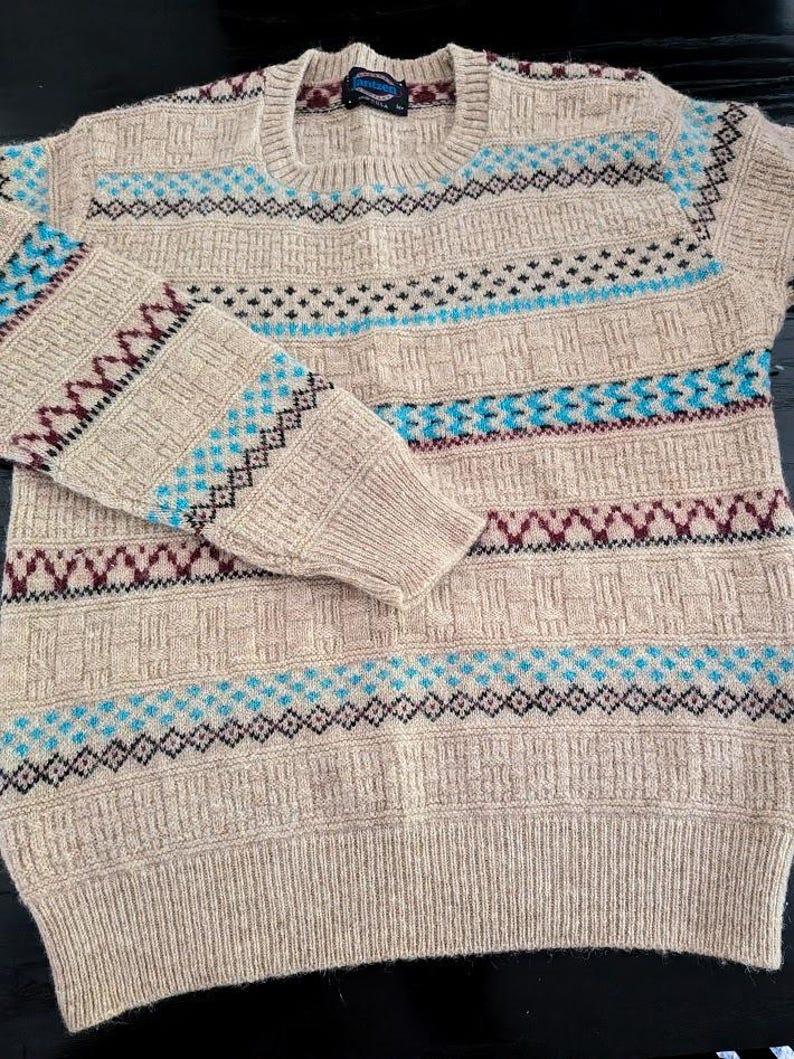 May include: A beige long-sleeve crew neck sweater featuring a textured knit pattern with horizontal bands of geometric designs in bright blue and dark maroon. The sweater has ribbed cuffs, a ribbed hem, and a ribbed neckline with a small interior label.