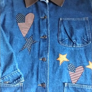 May include: A blue denim jacket with a brown corduroy collar. The jacket has a heart-shaped patch with an American flag design, a star-shaped patch, and a pocket on the front. The jacket has the word "HAK'S" on the collar.