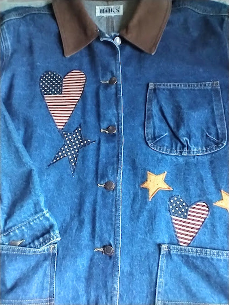 May include: A blue denim jacket with a brown corduroy collar. The jacket has a heart-shaped patch with an American flag design, a star-shaped patch, and a pocket on the front. The jacket has the word "HAK'S" on the collar.