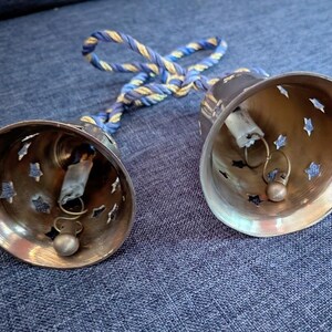 K&ouml;nnte beinhalten: Zwei Messingglocken mit sternf&ouml;rmigen Aussparungen und kleinen Kl&ouml;ppeln. Die Glocken sind durch ein blau-goldenes gedrehtes Seil verbunden. Die Glocken liegen auf einer blauen Jeansoberfl&auml;che.