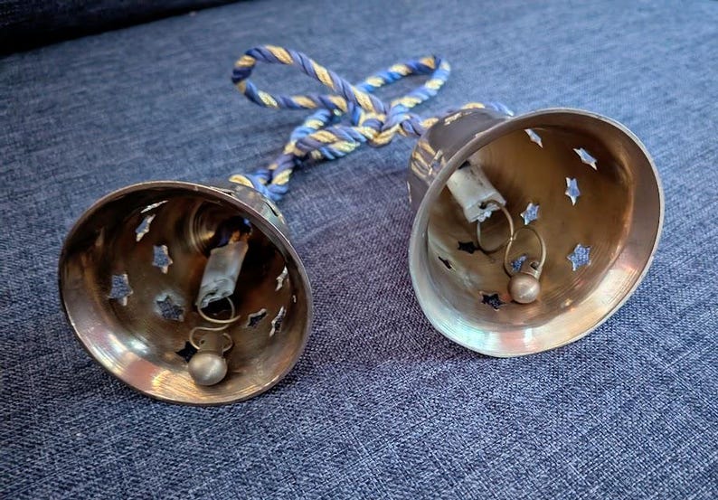 K&ouml;nnte beinhalten: Zwei Messingglocken mit sternf&ouml;rmigen Aussparungen und kleinen Kl&ouml;ppeln. Die Glocken sind durch ein blau-goldenes gedrehtes Seil verbunden. Die Glocken liegen auf einer blauen Jeansoberfl&auml;che.