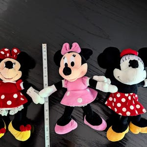 May include: Three plush Minnie Mouse dolls with black ears, white gloves, and yellow shoes. The left doll wears a red polka-dot dress, the centre doll wears a pink polka-dot dress, and the right doll wears a red polka-dot skirt. A ruler shows 28 cm.
