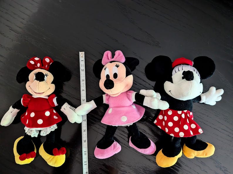 May include: Three plush Minnie Mouse dolls with black ears, white gloves, and yellow shoes. The left doll wears a red polka-dot dress, the centre doll wears a pink polka-dot dress, and the right doll wears a red polka-dot skirt. A ruler shows 28 cm.