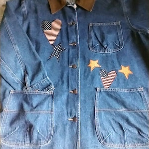 May include: A blue denim jacket with a brown corduroy collar. The jacket has a variety of patches, including hearts, stars, and a small American flag. The jacket has two large pockets on the front and a smaller pocket on the right side of the chest. The jacket has a button closure.