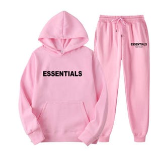 May include: A light pink hoodie and matching sweatpants. The hoodie features a front pocket and the word "ESSENTIALS" printed in black across the chest. The sweatpants have a drawstring waist and the word "ESSENTIALS" printed on the left leg.