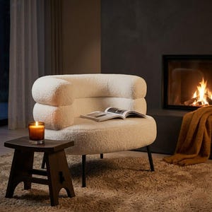 Puede incluir: Un sillón crema con un diseño redondeado y patas negras. Un libro abierto descansa sobre el asiento. Un pequeño taburete de madera sostiene una vela encendida. Una chimenea con fuego y una manta marrón completan la escena.