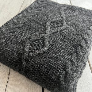 Puede incluir: Una manta de punto gris oscuro con un patrón de punto trenzado. La manta está doblada y colocada sobre una superficie de madera clara.