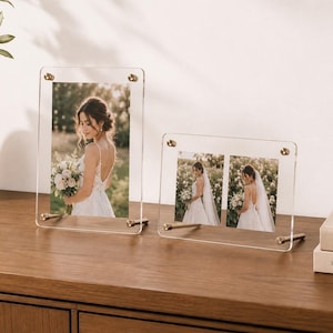 Peut inclure: Deux cadres photo en acrylique transparent avec des fixations en métal doré. Le cadre vertical contient une photo d'une personne en robe blanche, tandis que le cadre horizontal contient deux photos plus petites de la même personne en robe blanche.