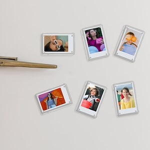 Peut inclure: Six cadres photo magnétiques rectangulaires en acrylique transparent fixés sur la porte d'un réfrigérateur blanc. Chaque cadre contient une petite photographie couleur montrant une personne différente dans divers décors extérieurs.